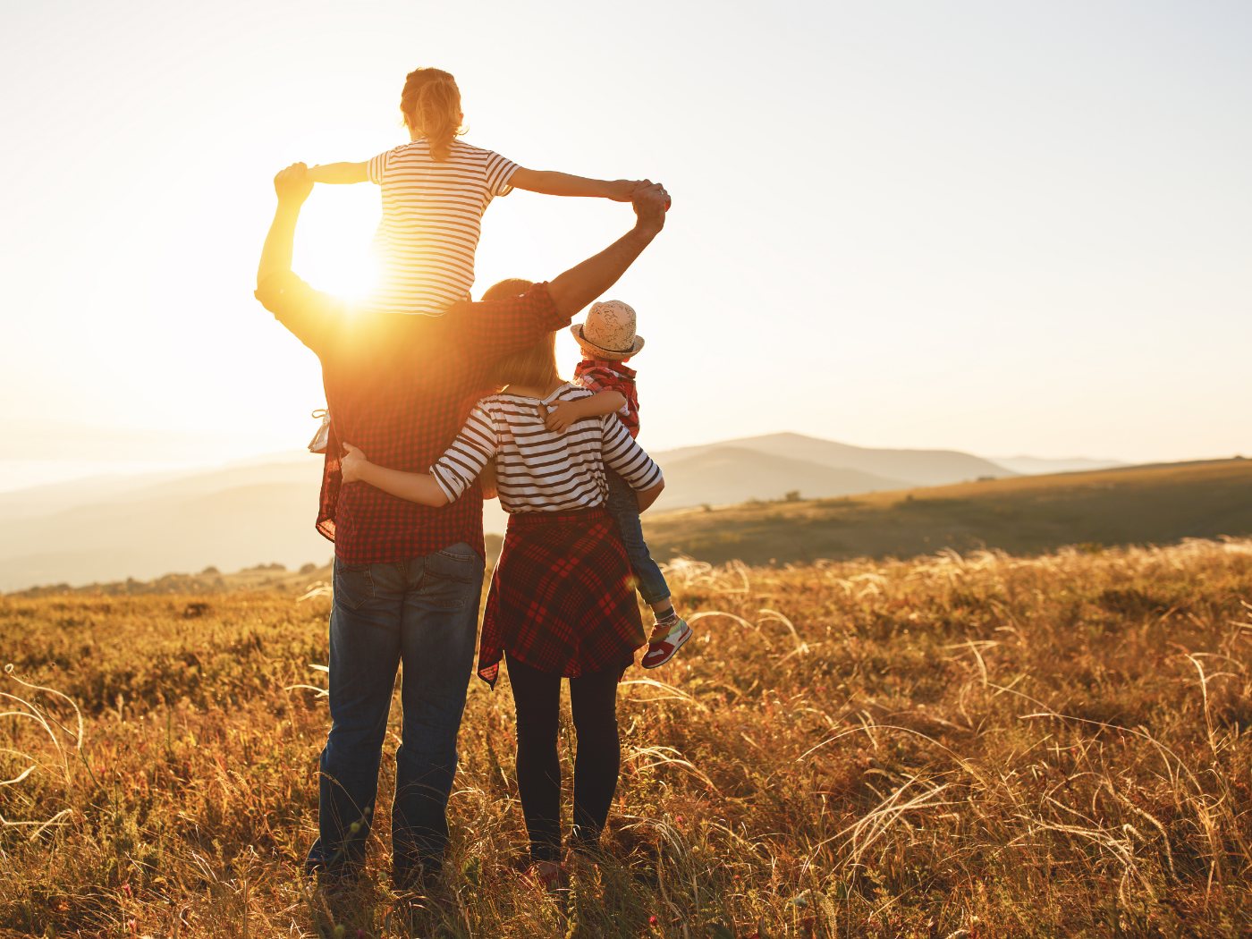 Vater trägt sein Kind auf den Schultern, die Familie geht durch ein Feld im Abendlicht